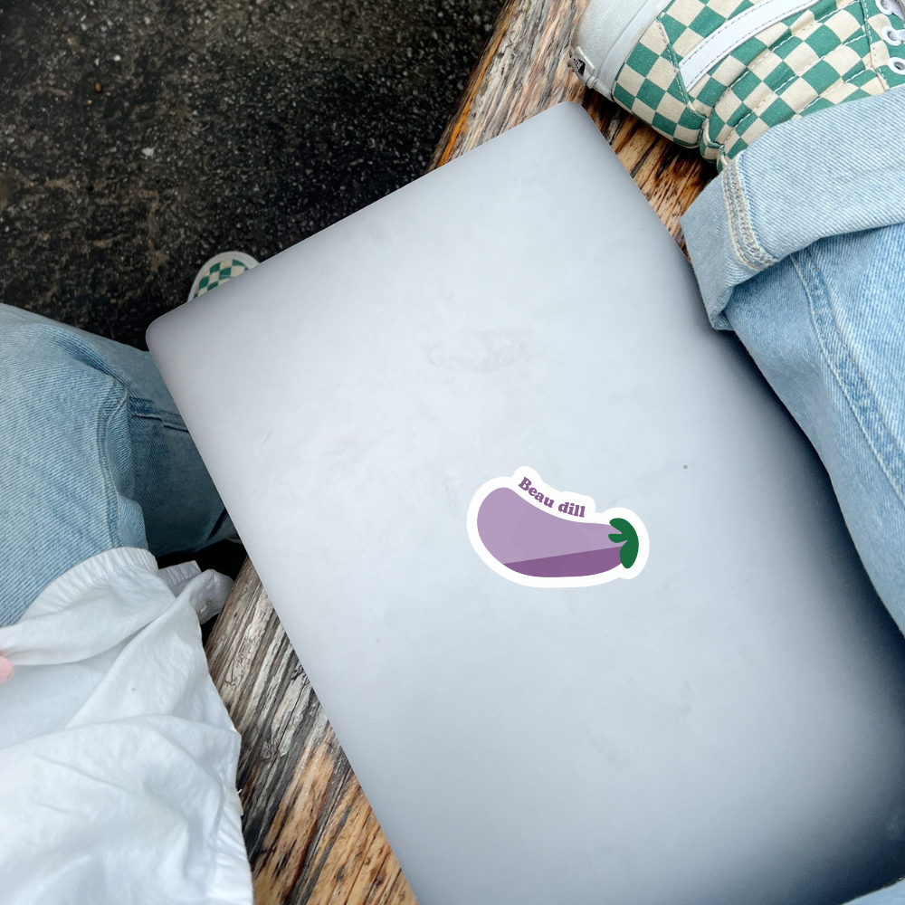 Mockup d'un autocollant érotique et ludique indiquant "Beau dill" avec une aubergine en emoji, collé sur un ordinateur portable entre les jambes d'une personne portant des jeans et des souliers de skateboard.