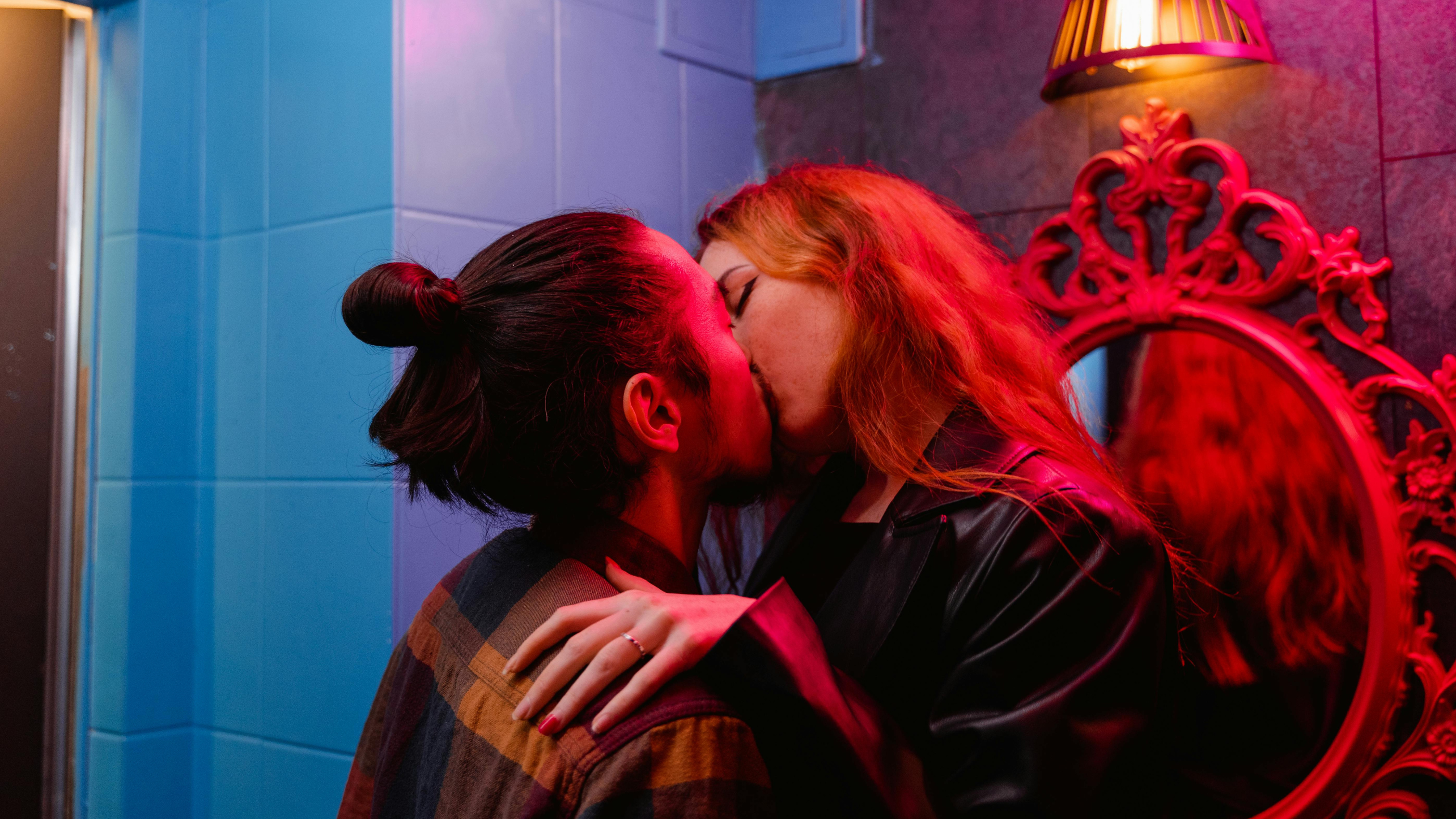 Photo représentant un couple de jeunes adultes qui s'embrasse passionnément contre un mur, dans une ambiance feutrée et intime. Peut-être un bar ou une soirée arrosée. Intime, sexy, coquin, érotique, spicy.